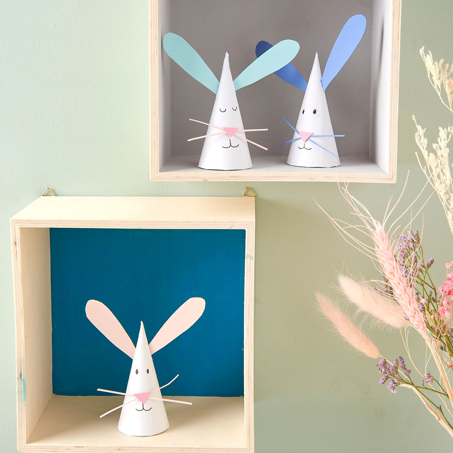 Bastelanleitung Osterhasen aus Spitztüten: Zwei weiße Papierhasen mit blauen und rosa Ohren in Holzkästen.