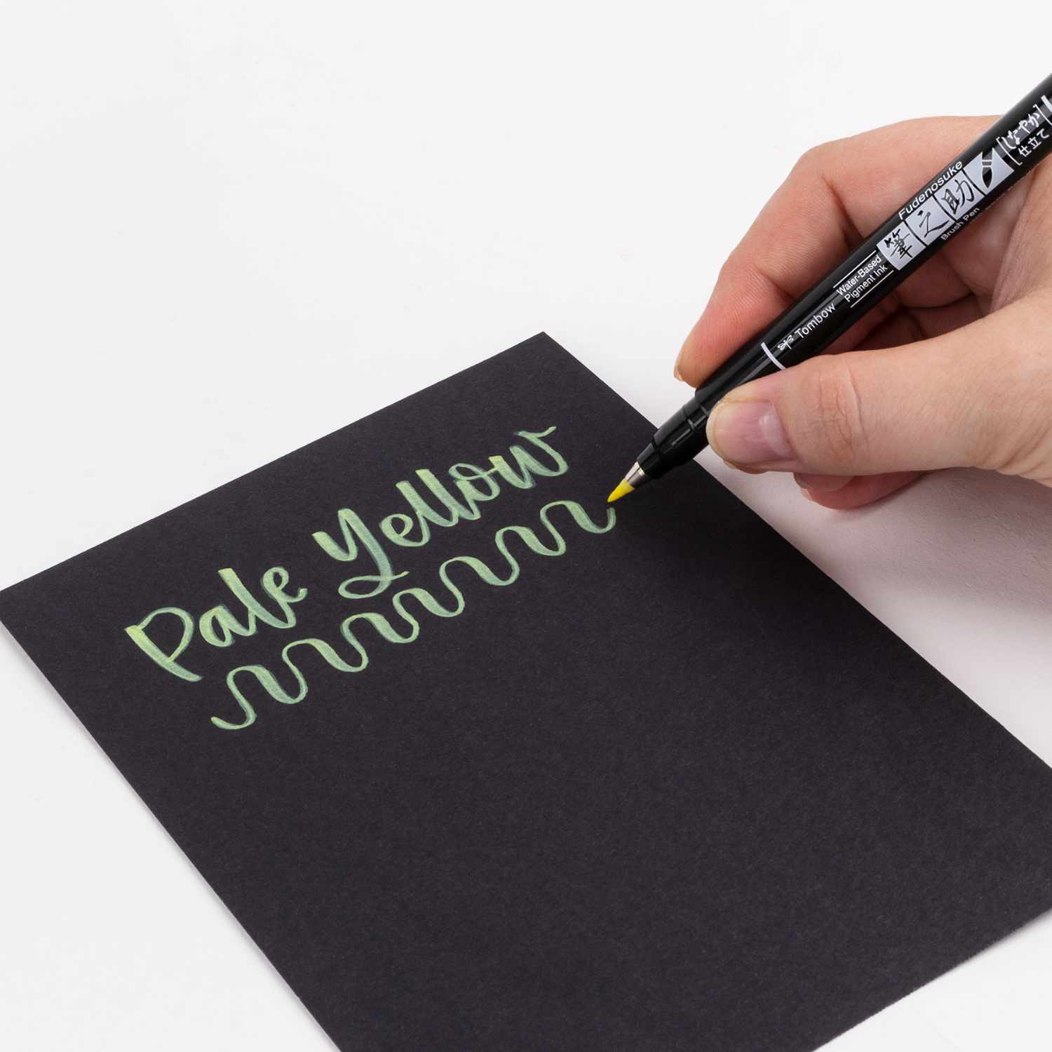 Fudenosuke Brush Pen Pastel schreibt in blassem Gelb auf schwarzem Papier. Hand hält Tombow-Stift.