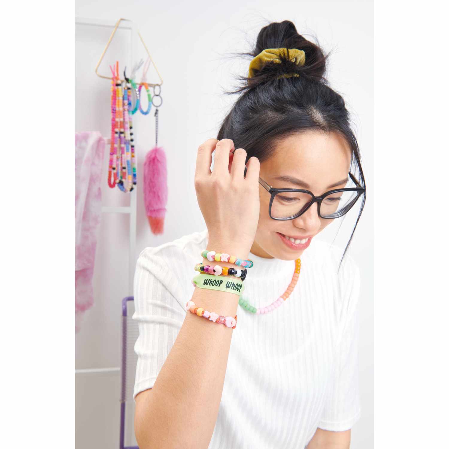 itoshii - Ponii Beads Bügelmotiv Slogans: Frau mit bunten Armbändern, lächelnd, vor Bastelzubehör.