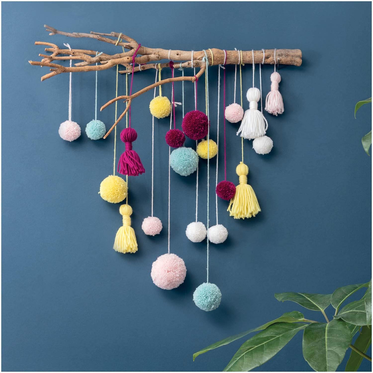 Bastelanleitung Wandbehang aus Pompons und Tasseln: Bunte Pompons und Tasseln an einem Ast vor blauem Hintergrund.