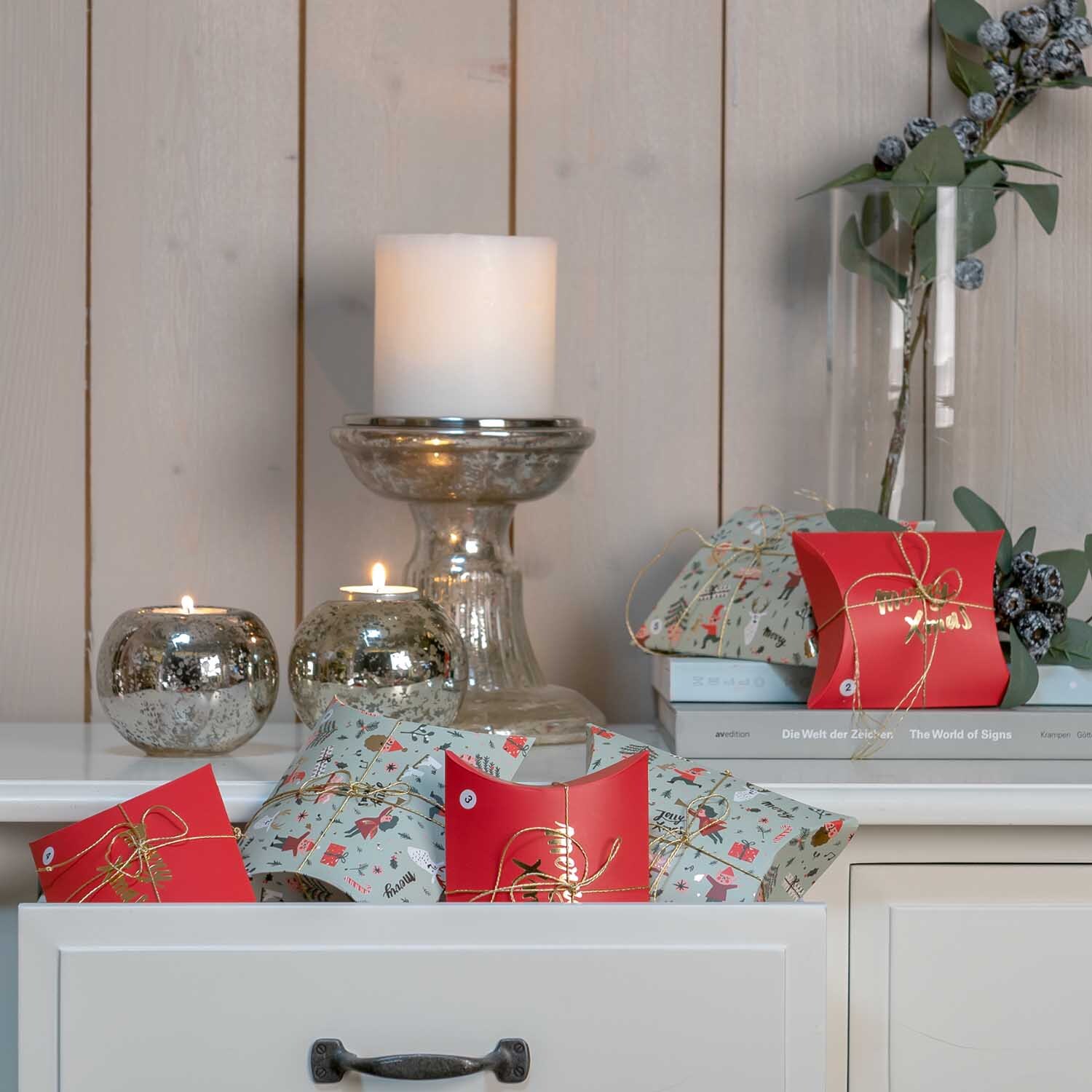 Paper Poetry Geschenkschachteln Jolly Christmas Classic, dekoriert mit Kerzen und Zweigen auf einem weißen Schrank, ideal für festliche Anlässe.