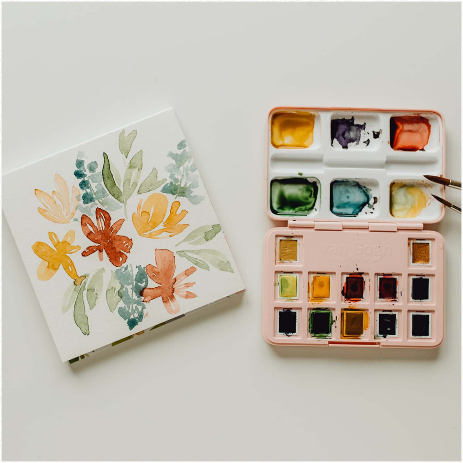 van Gogh Aquarellpapier 15x15cm mit floralen Motiven neben einem Aquarellkasten in Pastelltönen. Ideal für kreative Projekte.