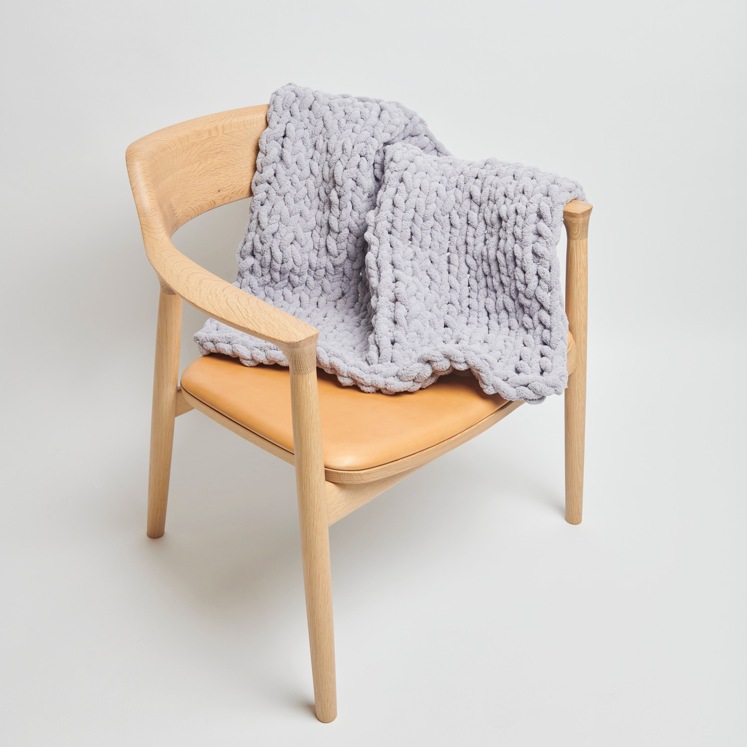 Chenillove Super Big: Weiche, lavendelfarbene Strickdecke auf einem modernen Holzstuhl drapiert.