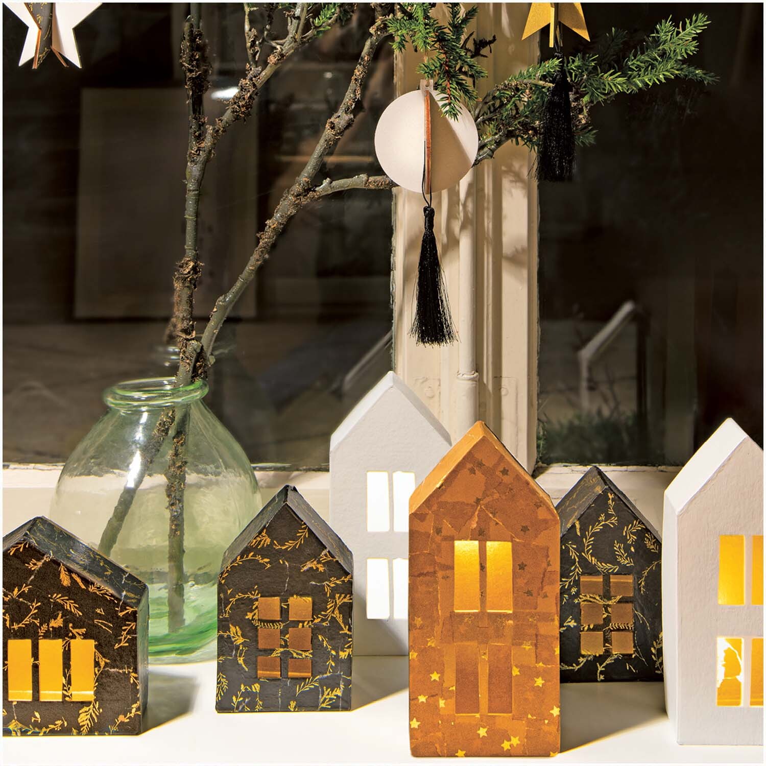 Bastelanleitung Weihnachtsdeko mit Paper Patch Papieren: Dekorative Papierhäuser in festlichen Farben vor einem Fenster mit Zweigen und Ornamenten.
