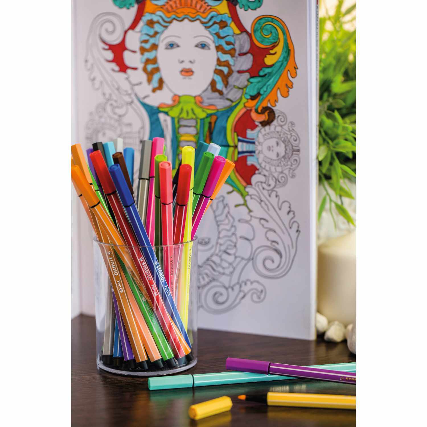 Pen 68 im Etui: Bunte Stifte in einem Glas vor einem farbenfrohen Malbuch, ideal für kreative Projekte.