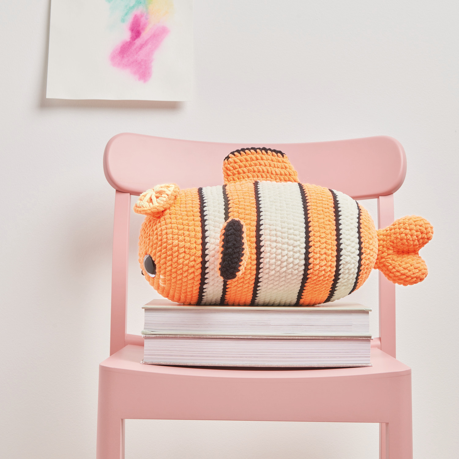 Häkelset Crazy Clown Fish Modell 04: Orange-weißer gehäkelter Fisch auf rosa Stuhl, dekorativ platziert.