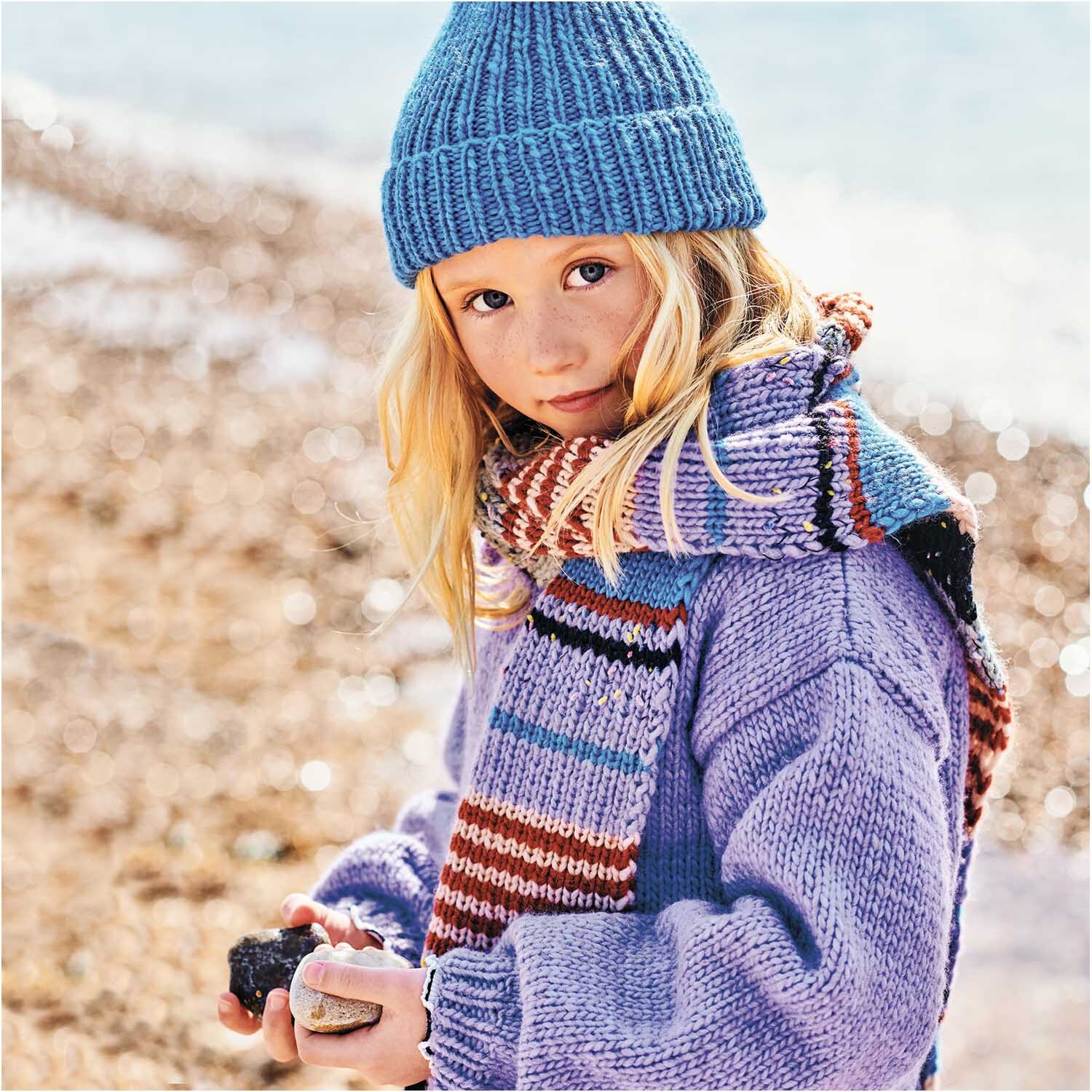 Essentials Super aran: Kind am Strand in lila Pullover und gestreiftem Schal, trägt blaue Mütze.