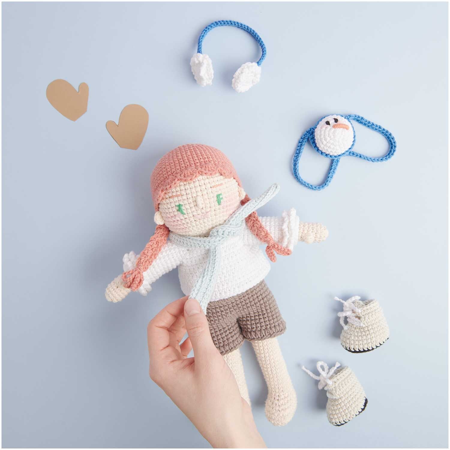 Creative Ricorumi dk: Hand hält gehäkelte Puppe mit Zubehör auf hellblauem Hintergrund, von Rico Design.