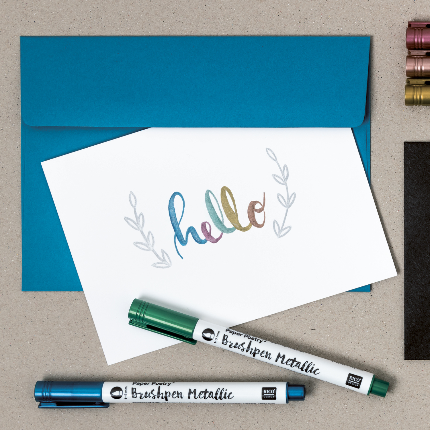 Anleitung Brushlettering: Karte mit farbigem "hello" und zwei Brushpen Metallic Stiften vor blauem Umschlag.