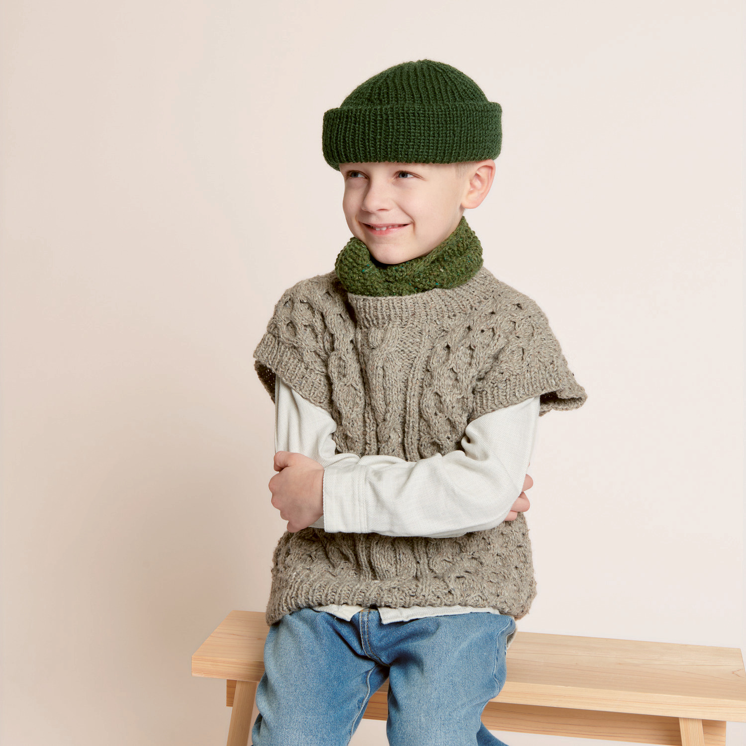 Strickset Kinderpullunder Modell 26: Kind trägt grauen Pullunder, grüne Mütze und Schal, sitzt auf Holzbank.