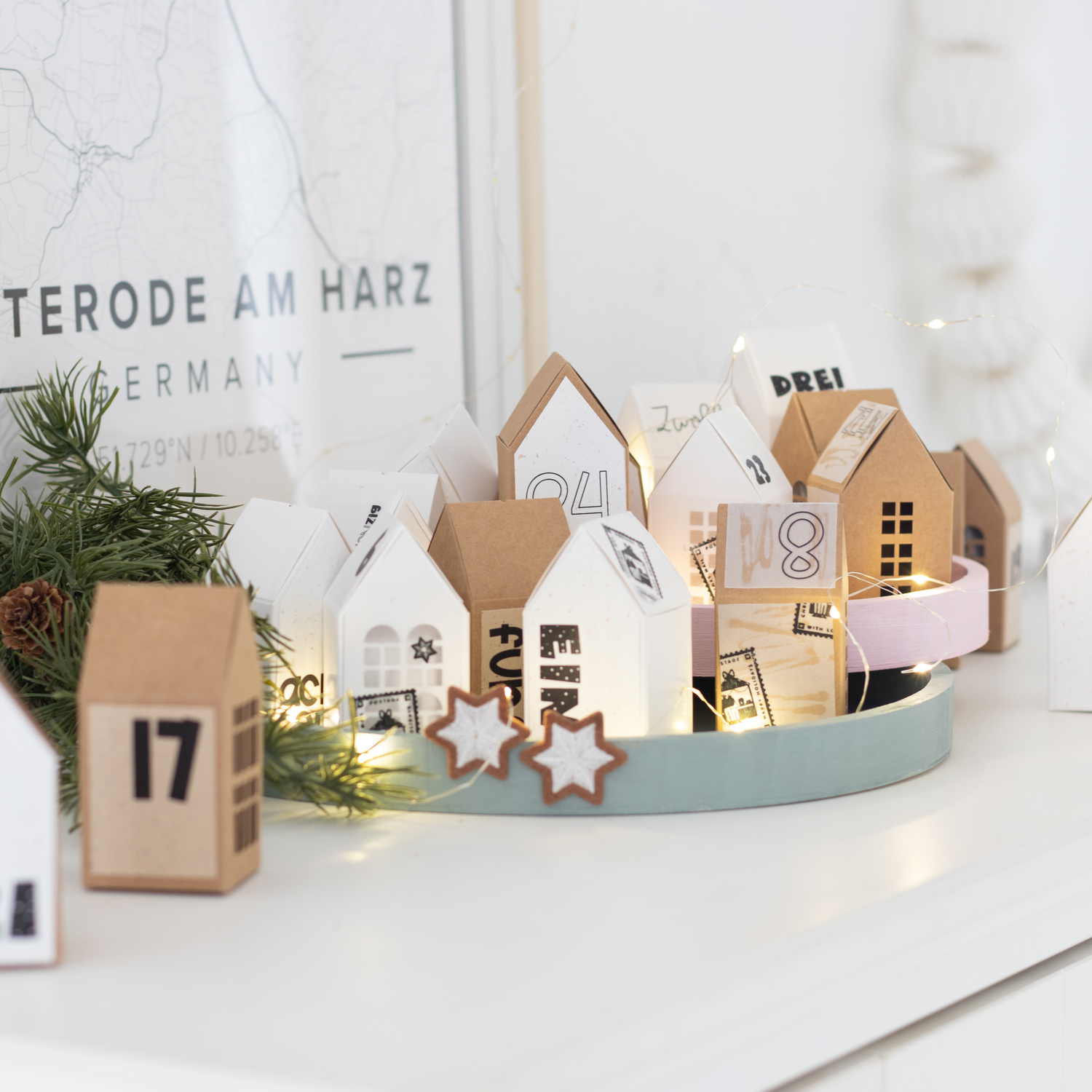 Inspiration Adventskalender Weihnachtsdorf: Dekoratives Dorf aus Papphäusern in Weiß und Braun auf einem Tablett, umgeben von Lichtern.