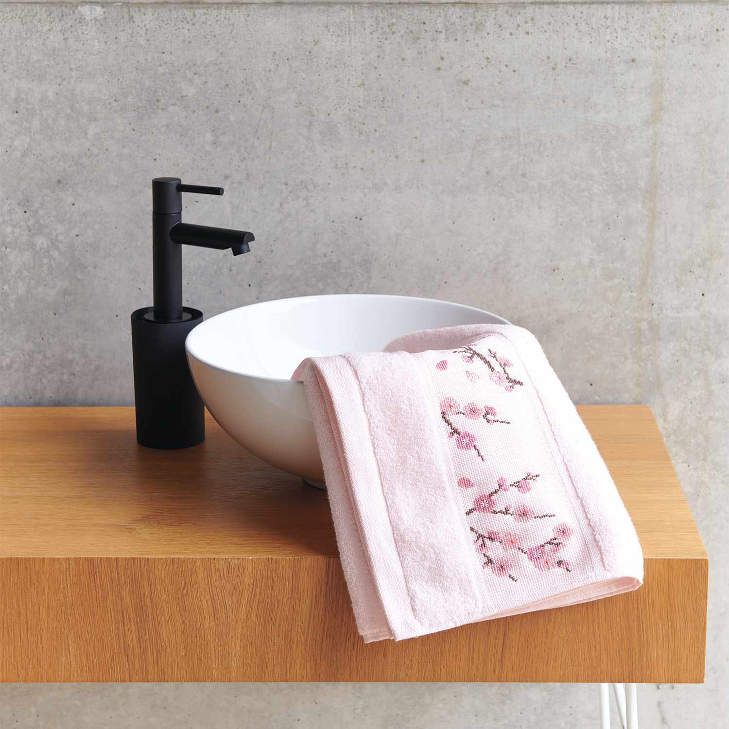 Stickanleitung Handtuch mit Kirschblüten-Zweigen auf Holzablage neben weißem Waschbecken und schwarzem Wasserhahn.