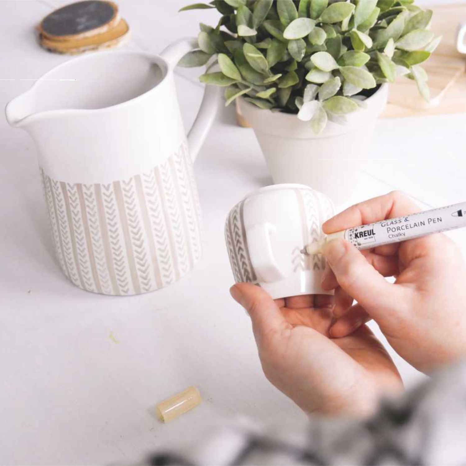 Glass & Porcelain Pen Chalky: Eine Hand verziert eine Tasse mit einem KREUL Stift, im Hintergrund Pflanzen und ein Krug.