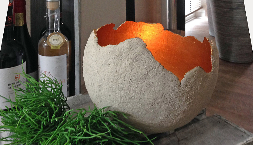 Bastelanleitung Deko Halbkugel aus Kreativbeton: Eine leuchtende, rustikale Betonschale auf einem Tisch, umgeben von Pflanzen und Flaschen.