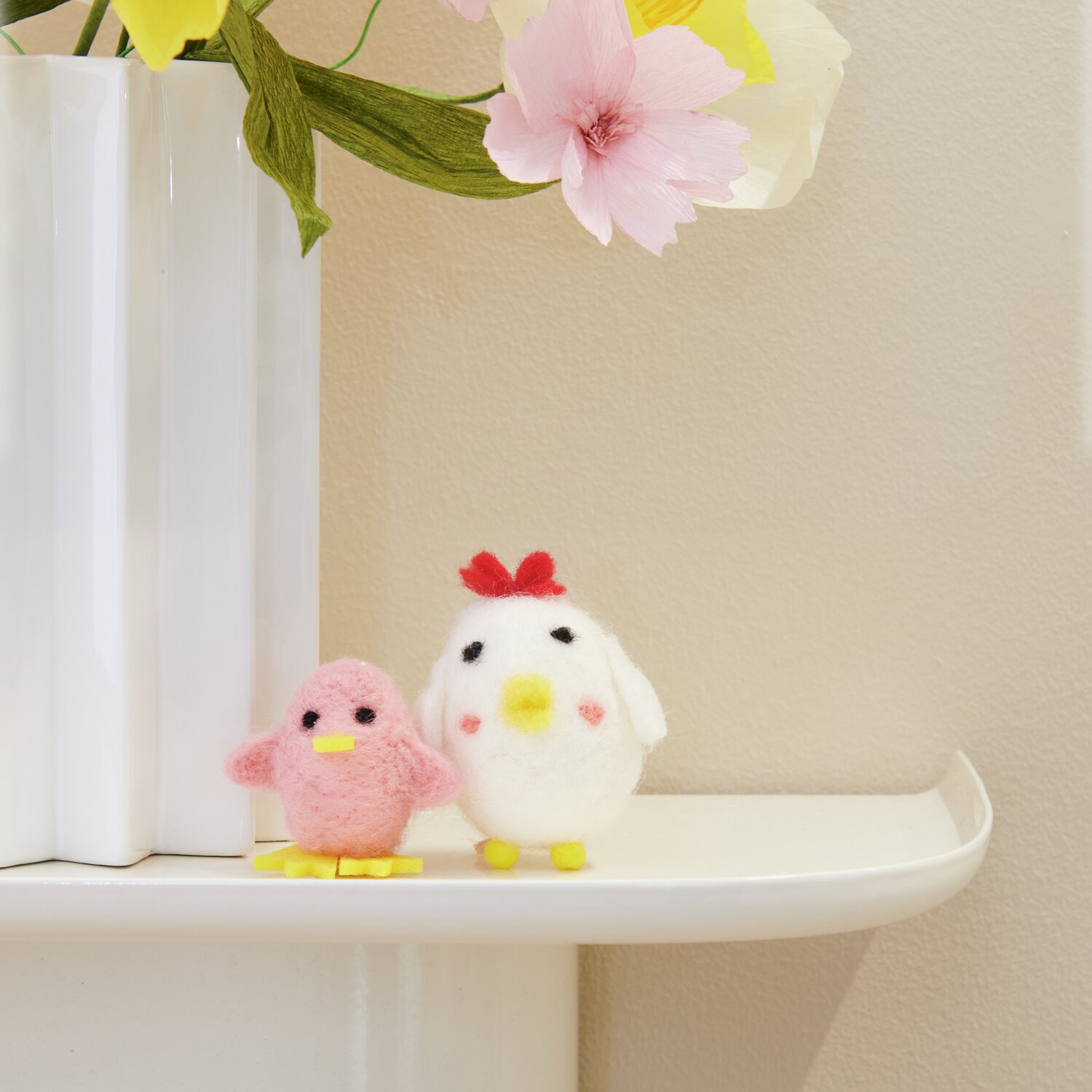Aufhänger Filz-Küken: Zwei Filzhänger in Kükenform, ein rosa und ein weißes, dekorieren ein Regal vor Blumen.