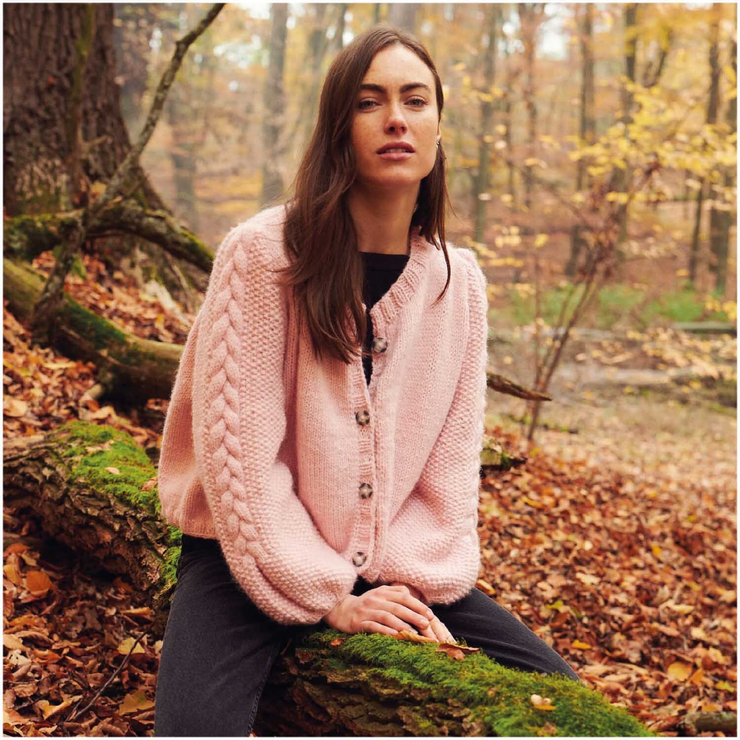 Essentials Alpaca Twist Chunky: Frau in rosa Strickjacke von Rico Design im herbstlichen Wald auf Baumstamm sitzend.