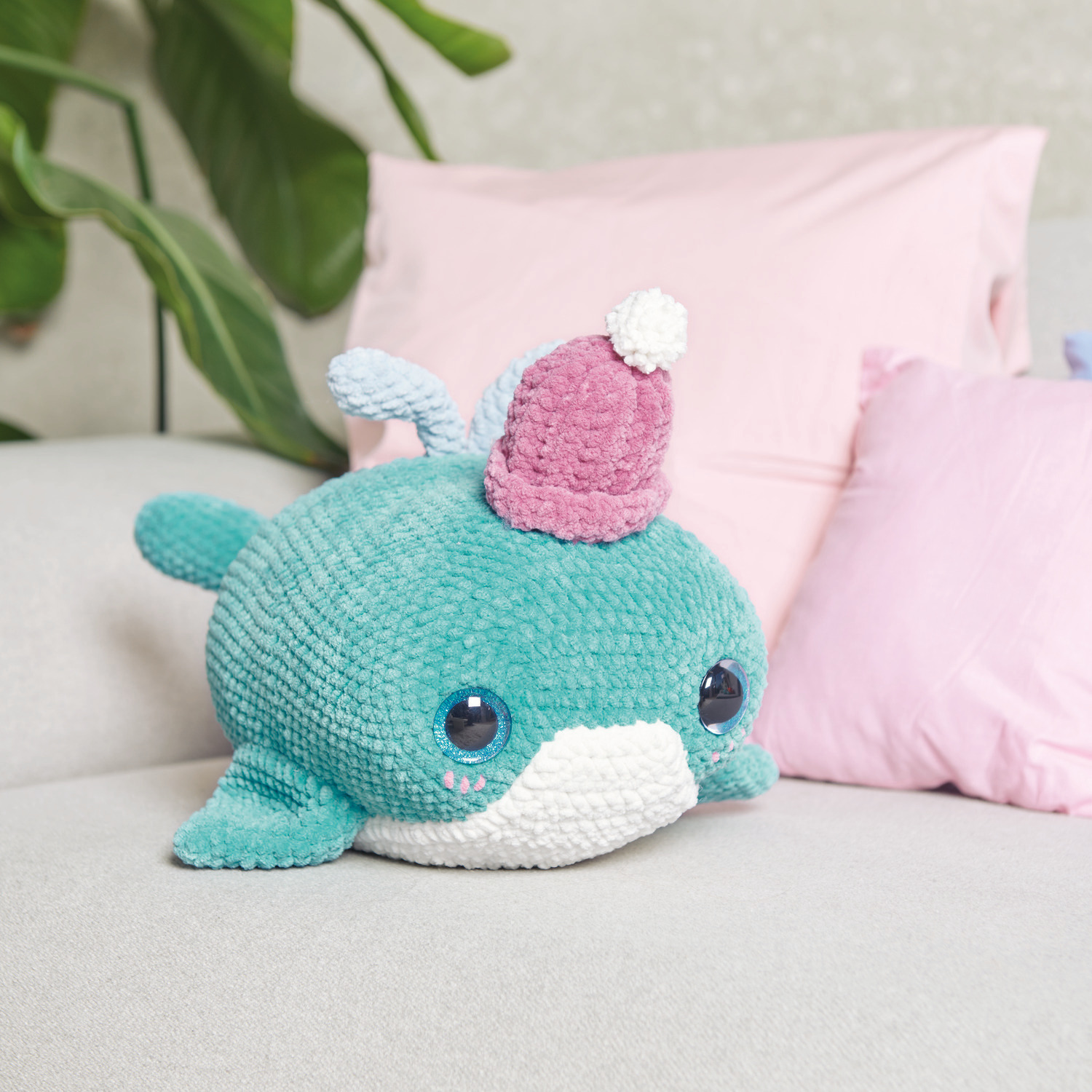 Häkelset Grandpa Whale Modell 06: Ein türkisfarbener Wal mit rosa Mütze auf einem Sofa.