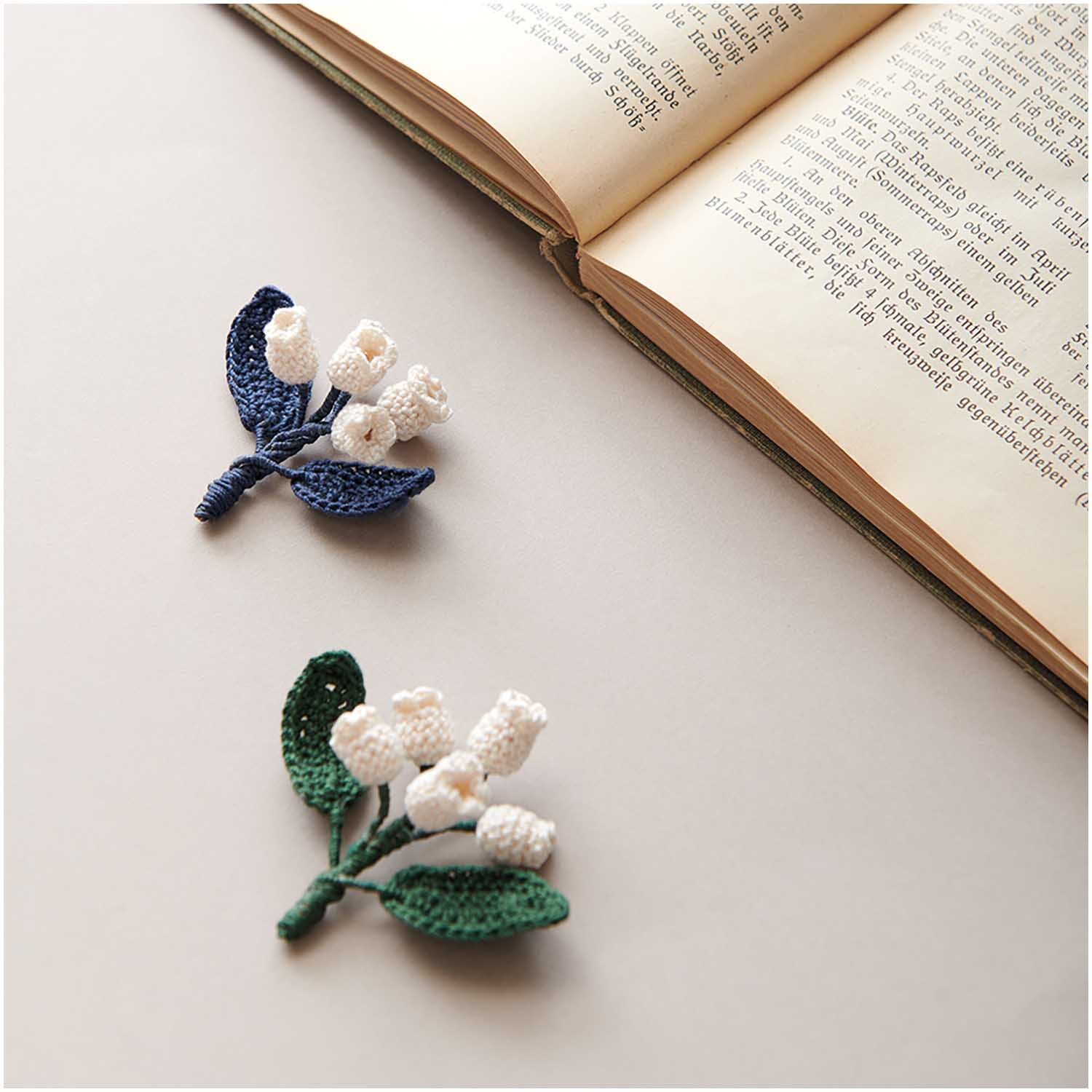 Essentials Crochet: Zwei gehäkelte Blumen neben einem aufgeschlagenen Buch, ideal für kreative Projekte.