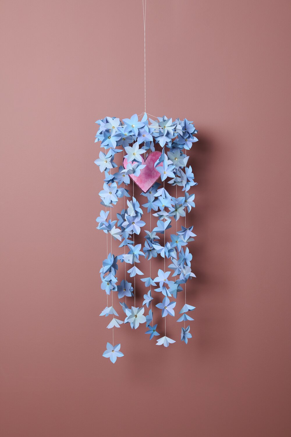 Papierblüten Bastelset La Vie en Rose: Blaue Papierblüten hängen dekorativ vor rosa Hintergrund.