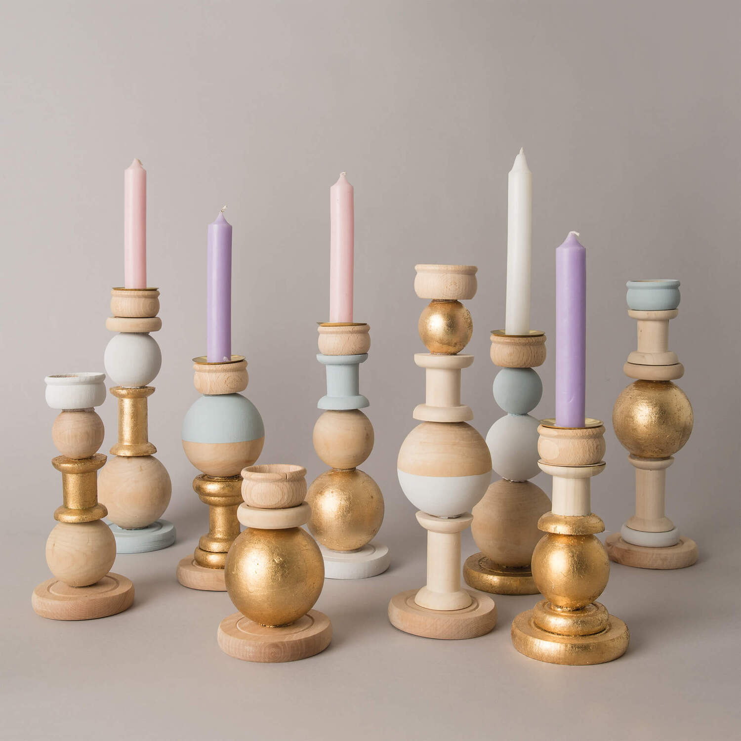 Bastelanleitung Kerzenhalter aus Holz: Verschiedene Holzkerzenhalter in Pastell- und Goldtönen mit Kerzen in Rosa, Weiß und Lila auf grauem Hintergrund.
