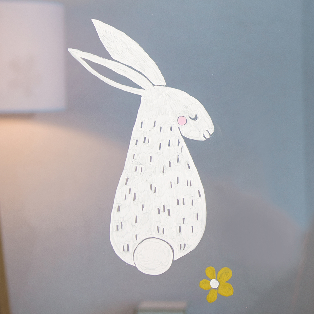 Anleitung Chalk Window Art Ostern: Ein weißer Hase mit rosa Wangen und gelber Blume auf Glas gemalt.