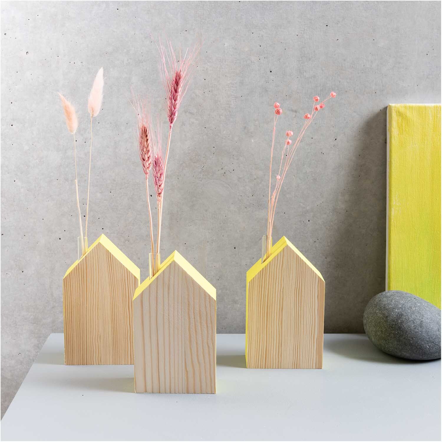 Holz-Haus mit kleiner Glasvase 9x15x5cm: Drei gelbe Holzvasen in Hausform mit Trockenblumen auf grauem Tisch.