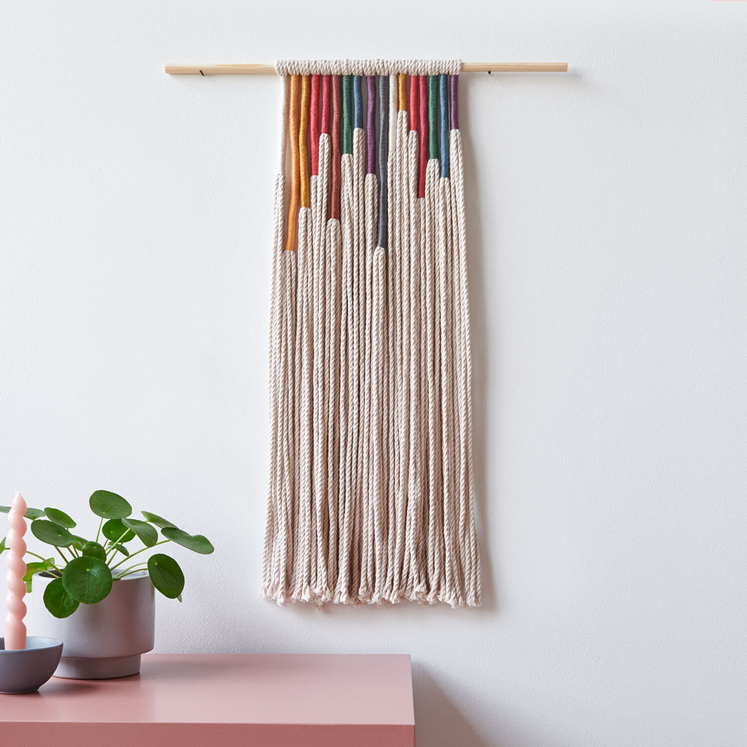 Bastelanleitung Sticktwist-Wanddeko Earthy: Bunte Fäden an Holzstab, dekorativ an weißer Wand.