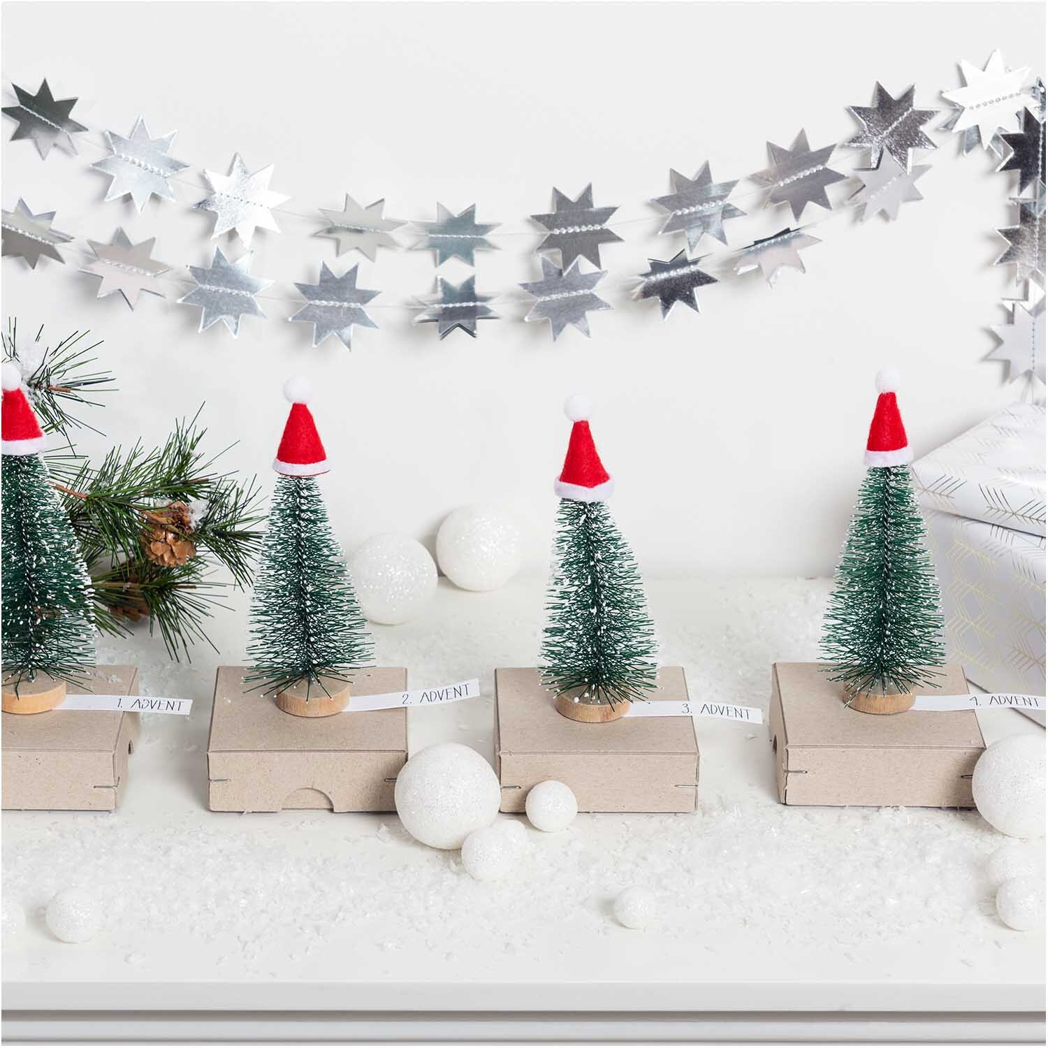Mini-Weihnachtsmützen rot 2x3,5cm 12 Stück: Vier Mini-Tannenbäume mit roten Mützen auf Kartons, umgeben von Schneebällen und silberner Sternengirlande.