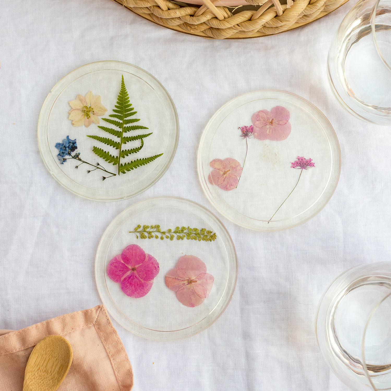 Bastelanleitung Untersetzer aus Resin mit gepressten Blumen, zeigt drei runde Untersetzer mit bunten Blüten und Farnen auf weißem Tuch.