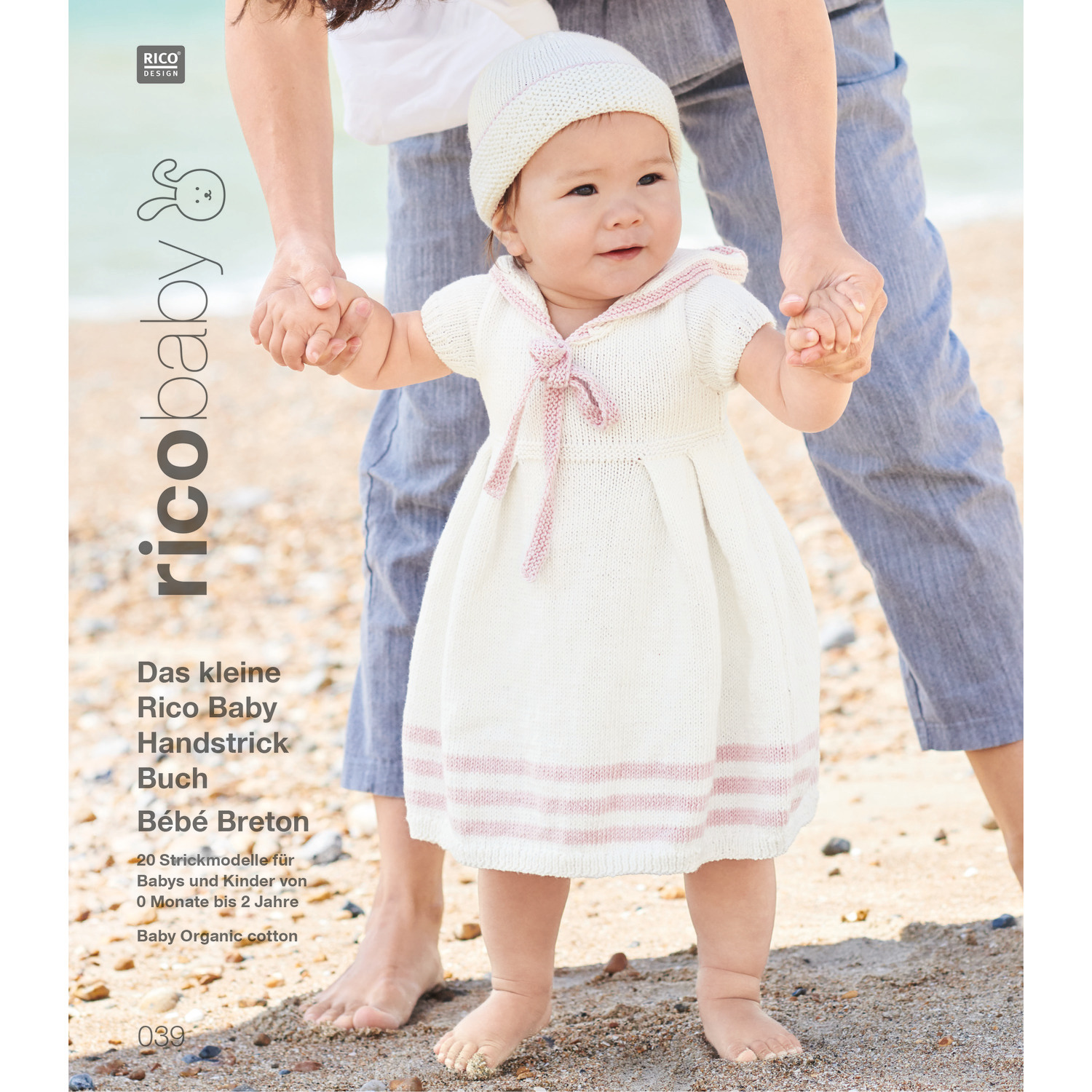 Rico Baby 039: Kleinkind in weißem Strickkleid mit rosa Streifen am Strand, gehalten von Erwachsenem.