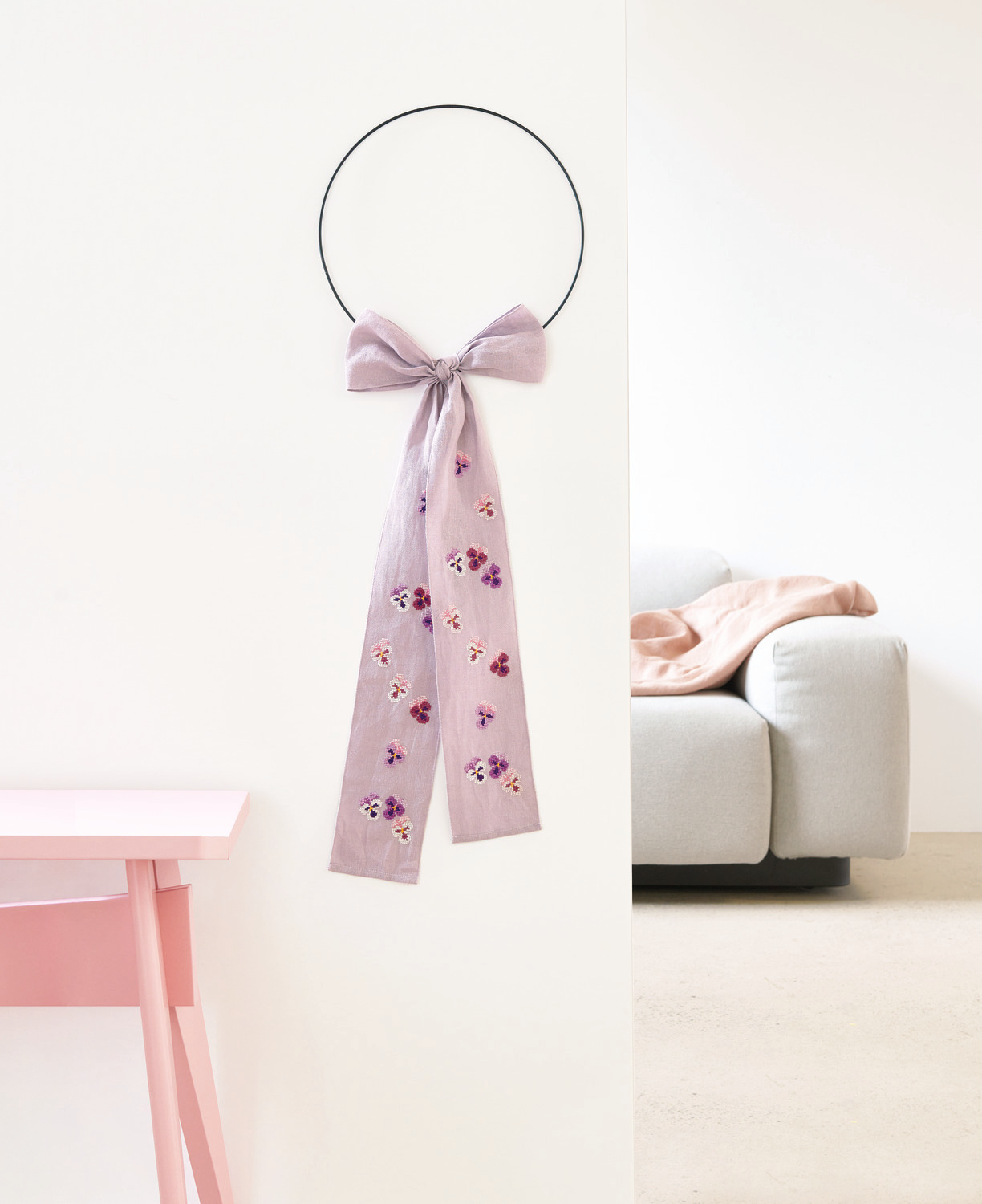 Stickpackung Schleife Stiefmütterchen von Rico Design: Elegante Schleife mit floralen Mustern an einem Metallring, dekorativ an einer Wand hängend.