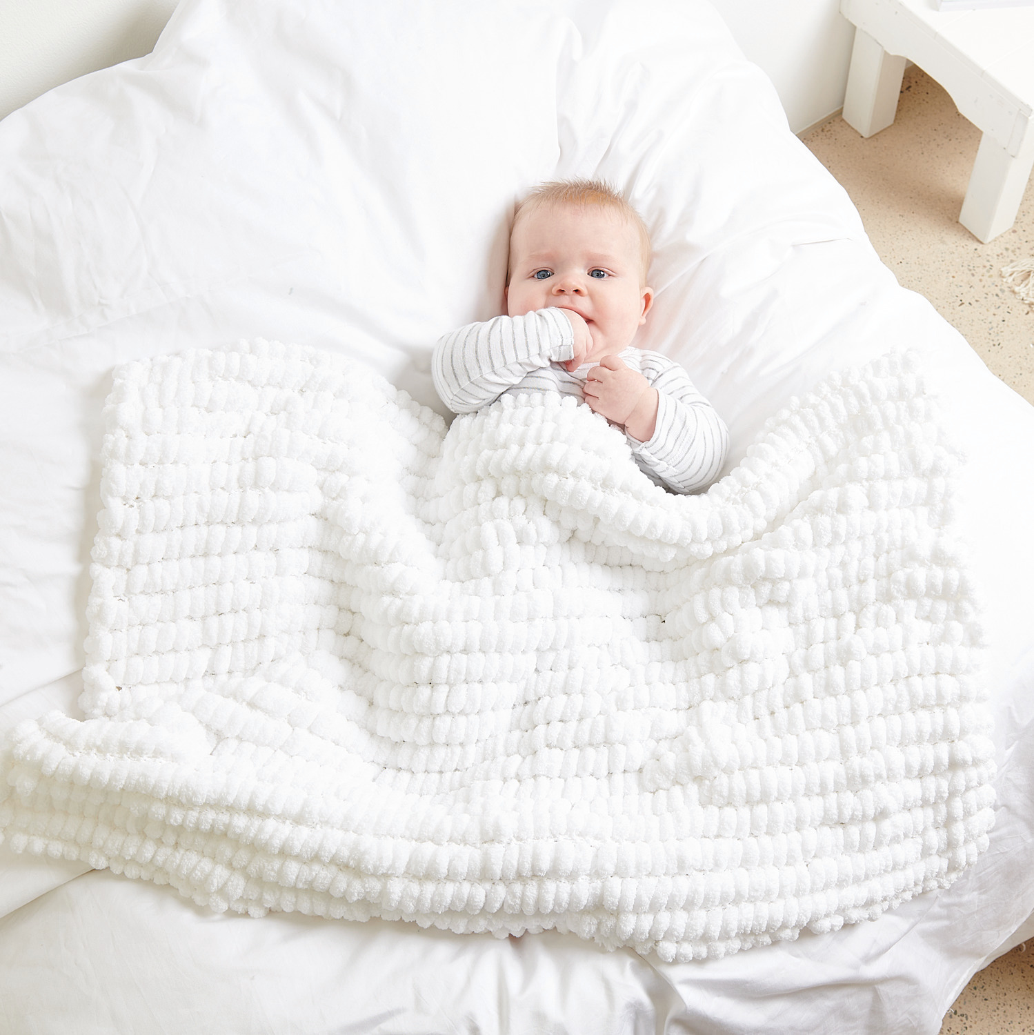 Pompon Babydecke in Weiß von Rico Design, kuschelig auf einem Bett liegend, 200g 100m.