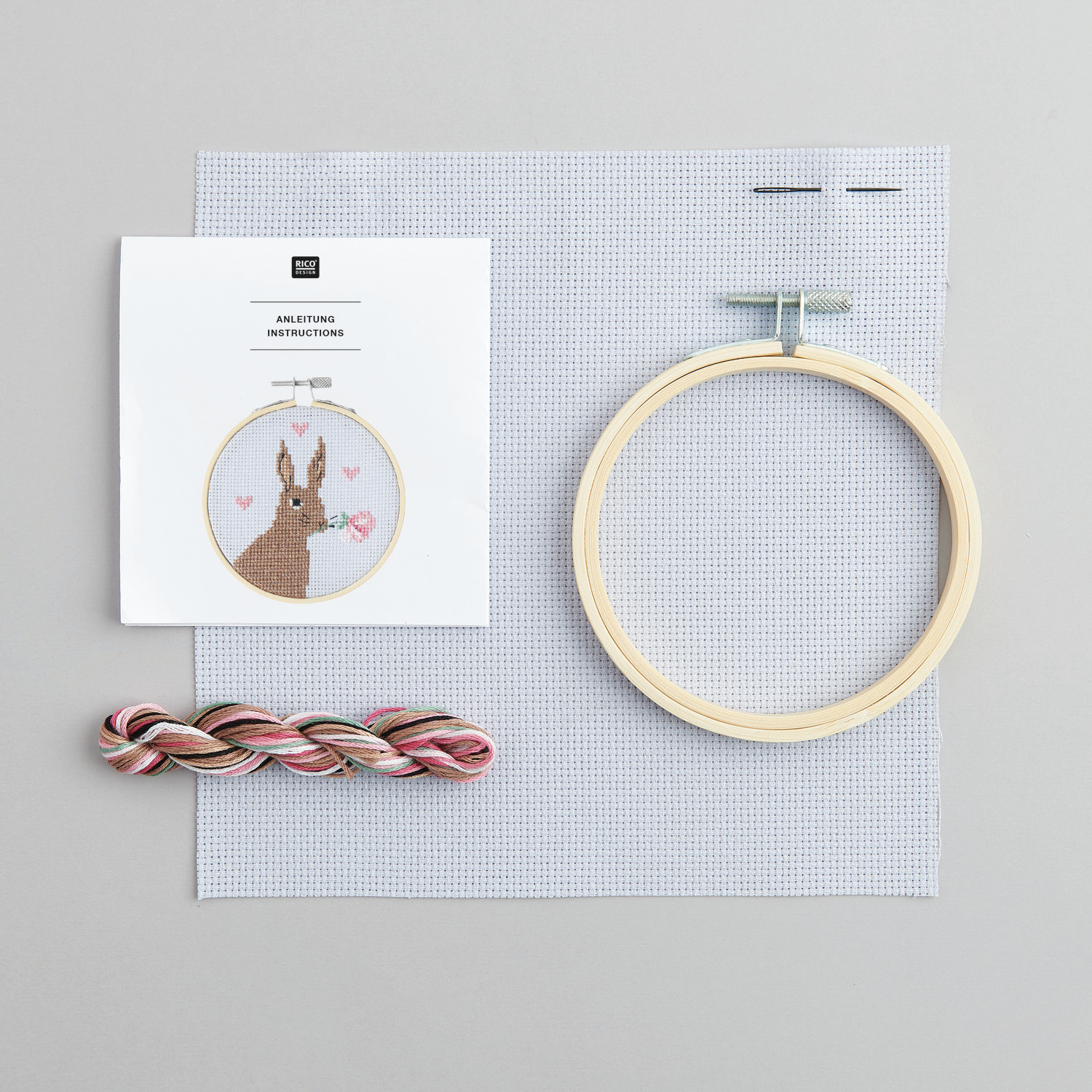 Stickpackung Bild Romantischer Hase zeigt einen Stickrahmen, Garn und Anleitung von Rico Design auf grauem Hintergrund.