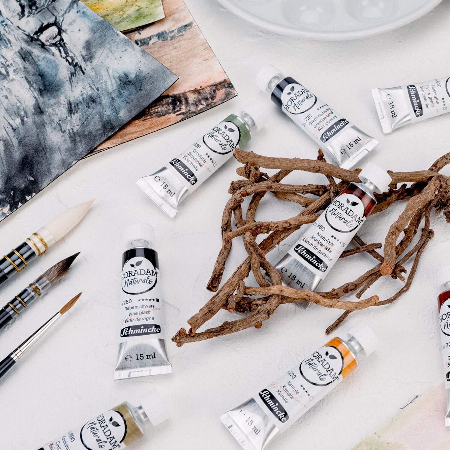 Horadam Naturals Aquarellfarben von Schmincke, 15ml Tuben, arrangiert mit Pinseln und Zweigen auf einem Tisch. Ideal für Künstler.