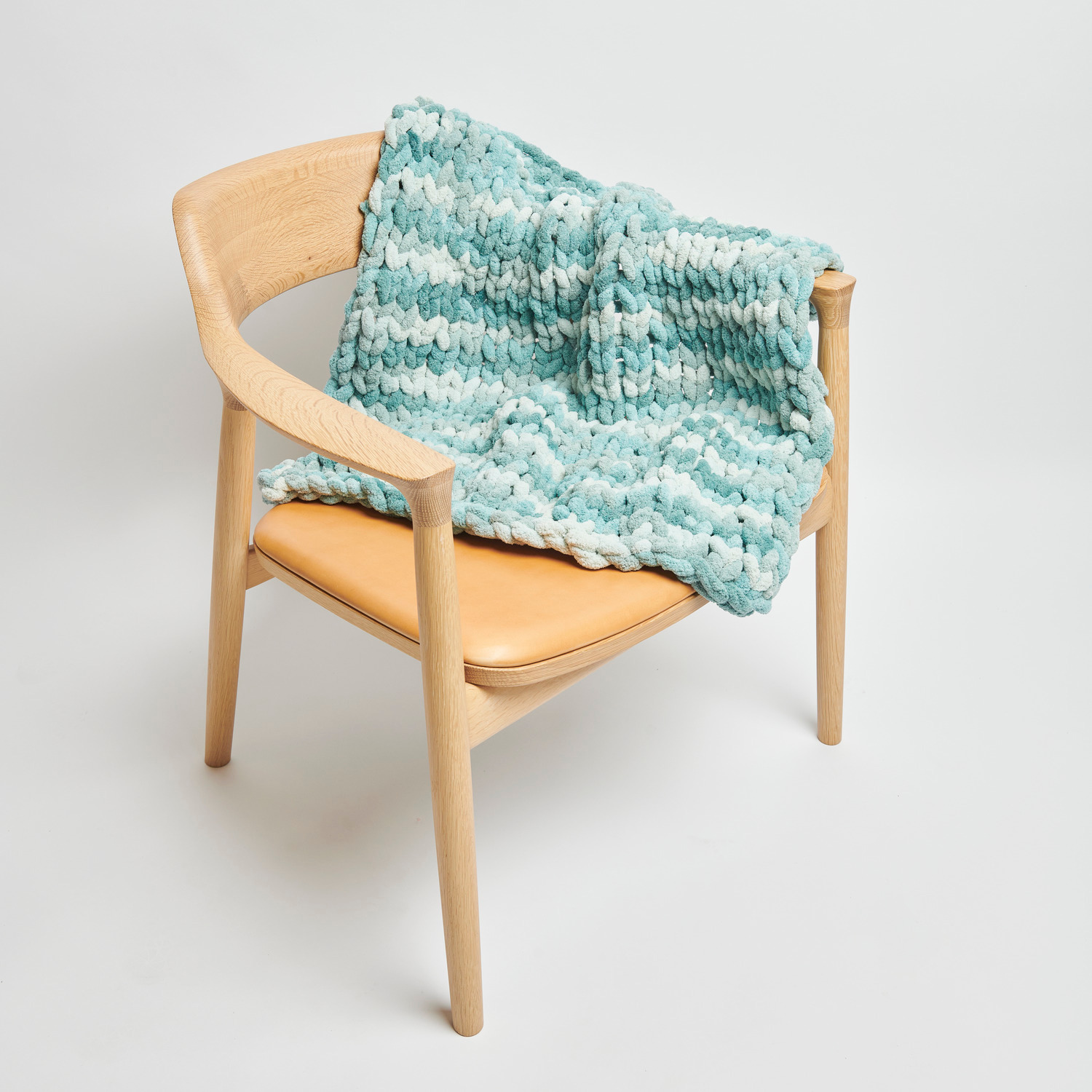 Chenillove Super Big Print: Weiche, aquafarbene Strickdecke von Rico Design auf einem Holzstuhl drapiert.