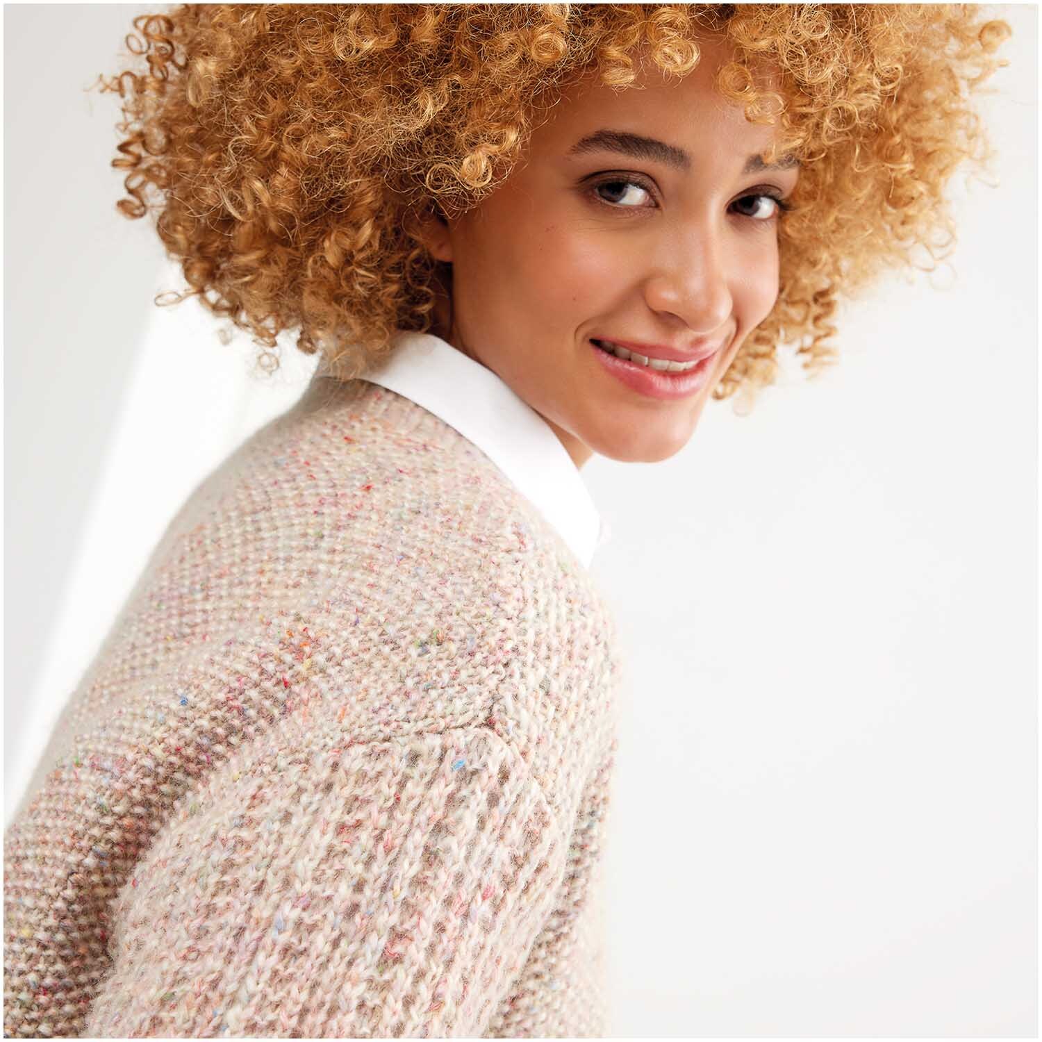 Strickset Pullover Modell 11: Frau mit lockigem Haar trägt einen gestrickten Pullover in sanften Tönen, lächelnd vor weißem Hintergrund.