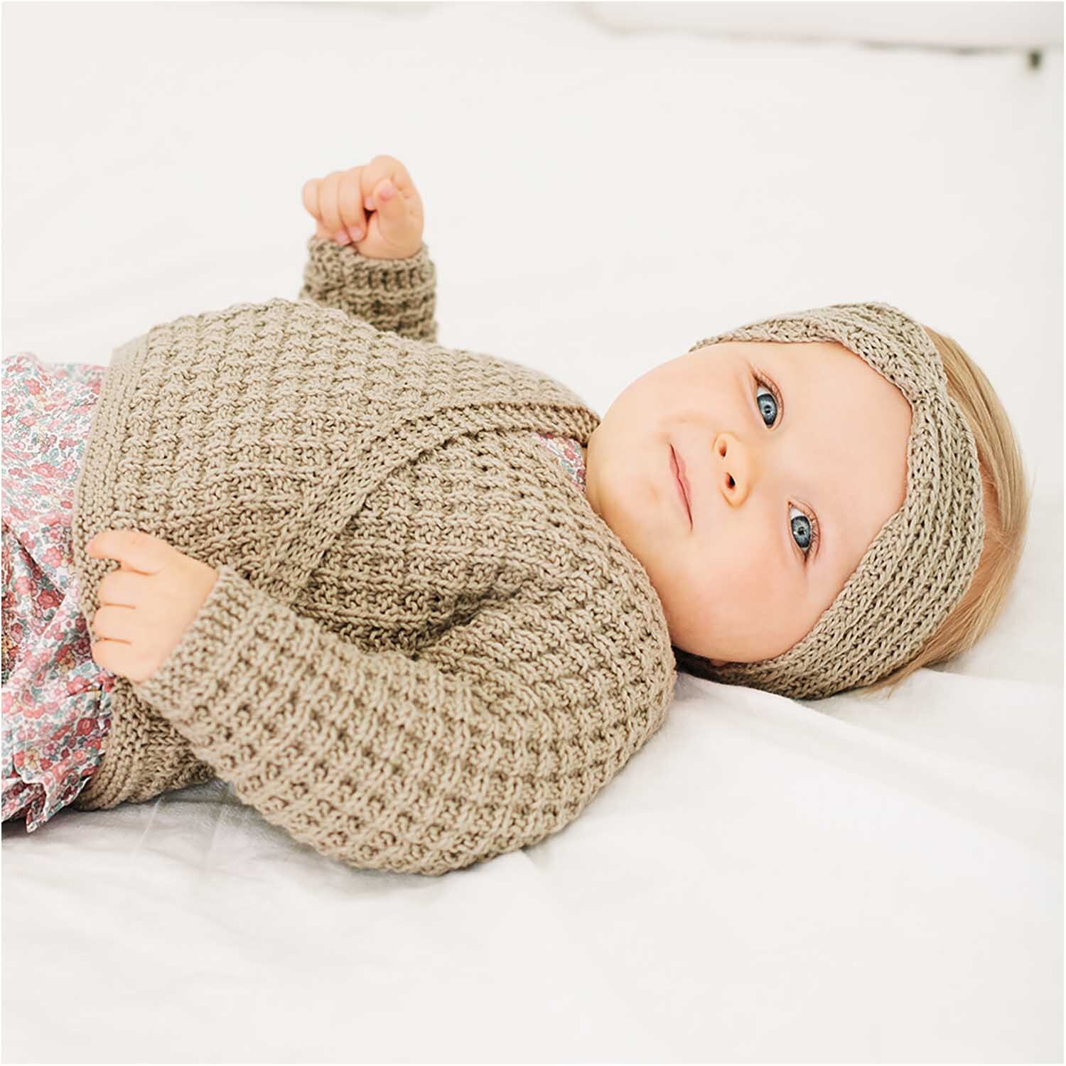 Rico Baby Cotton Soft dk: Ein Baby trägt einen gestrickten olivfarbenen Pullover und Stirnband auf einem weißen Bett.