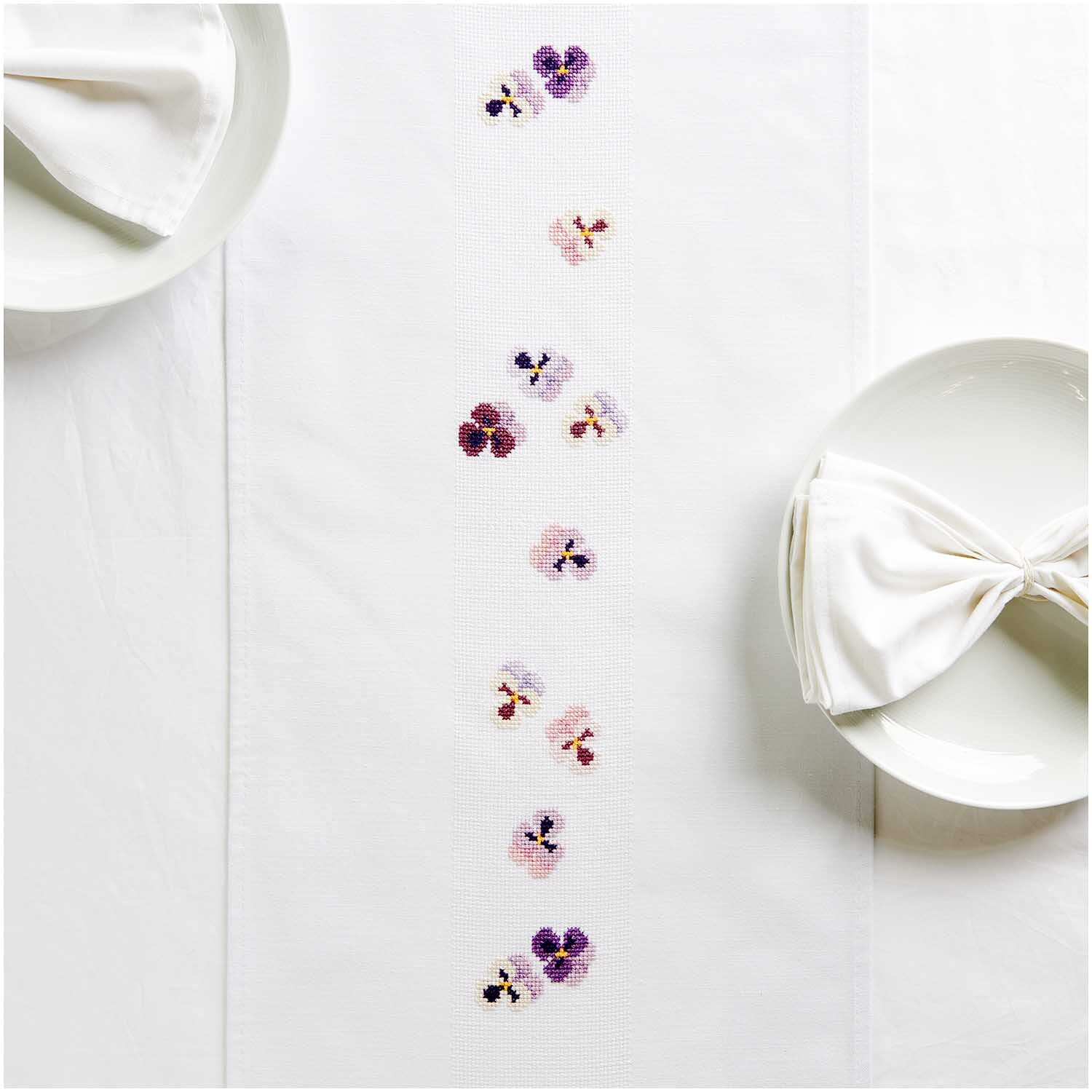 Stickpackung Tischband Stiefmütterchen von Rico Design, dekoriert mit zarten Blumenstickereien auf weißem Stoff, ideal für elegante Tischdekorationen.