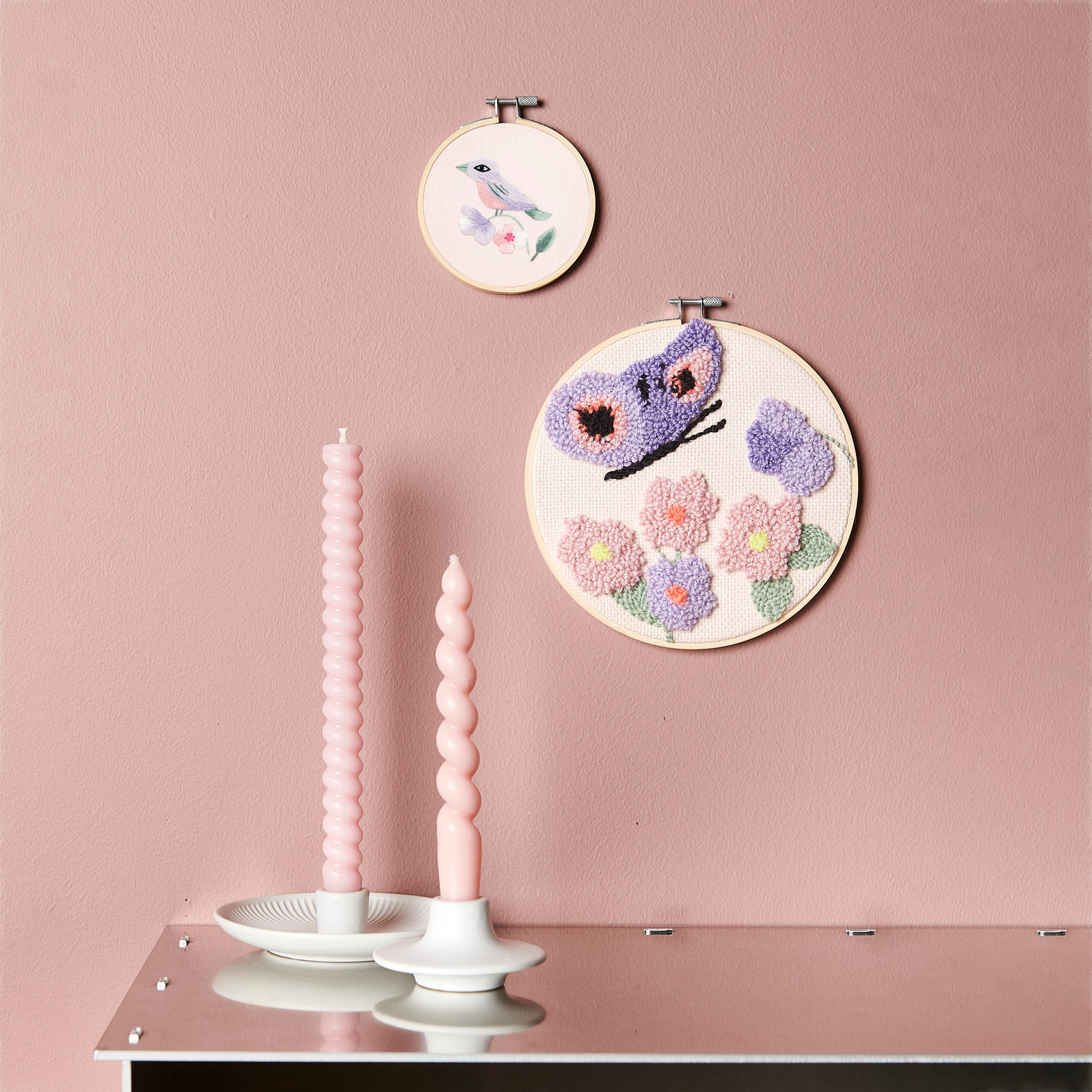 Stickpackung Vogel vorgezeichnet und Punch Needle Packung Schmetterling an rosa Wand, Ø10cm und Ø20cm, mit rosa Kerzen im Vordergrund.