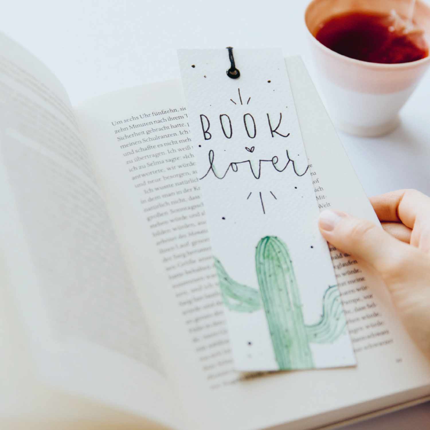 Anleitung Lesezeichen mit Kaktus: Ein Lesezeichen mit Kaktusmotiv liegt in einem offenen Buch neben einer Tasse Tee.