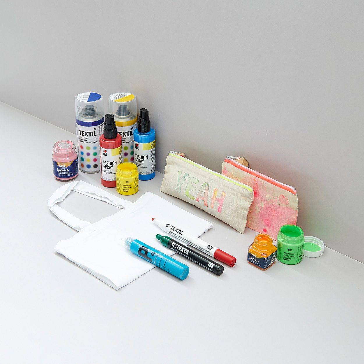 Stifte, Farben und Textilsprays in verschiedenen Farben und eine weiße Stofftasche gefaltet mit zwei Mäppchen. Fotosetting graue Ecke.