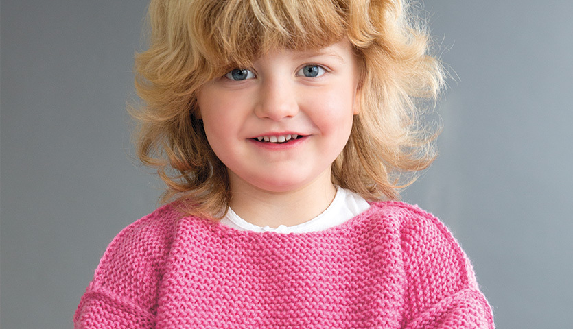 Strickanleitung Kinderpullover aus Soft Merino: Ein lächelndes Kind trägt einen rosa Pullover.