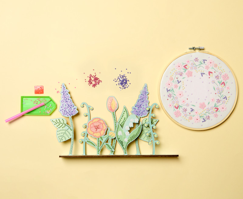 Ein florales Diamond-Painting auf einem Stickring und ein floraler Diamond-Painting-Dekoaufsteller, dazu das passende Zubehör. Flatlay von Produkten.