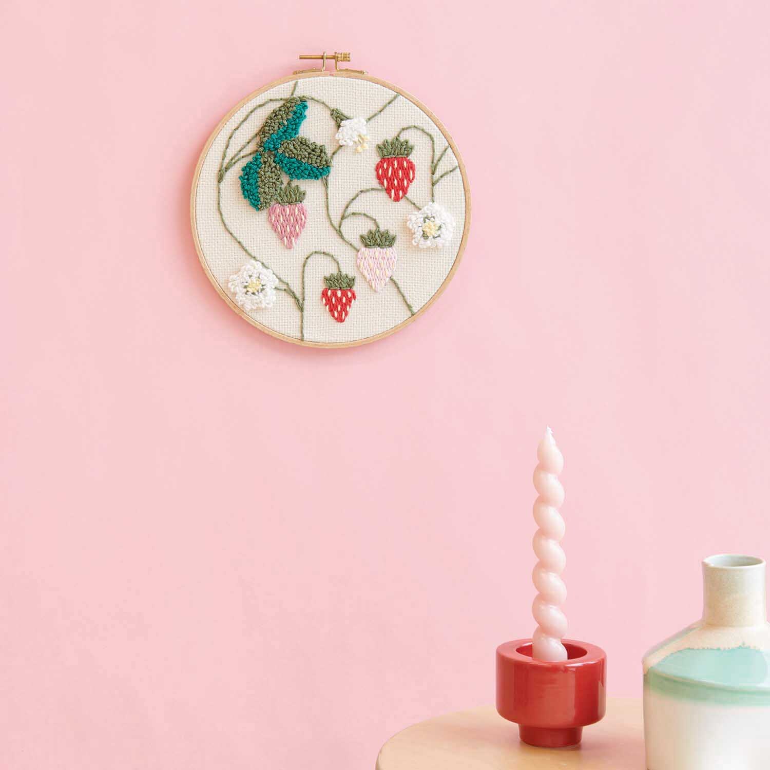 Anleitung Punch Needle Bild mit Erdbeeren gestalten: Wanddekoration mit Erdbeermotiv auf rosa Hintergrund, neben Kerzen und Vasen.