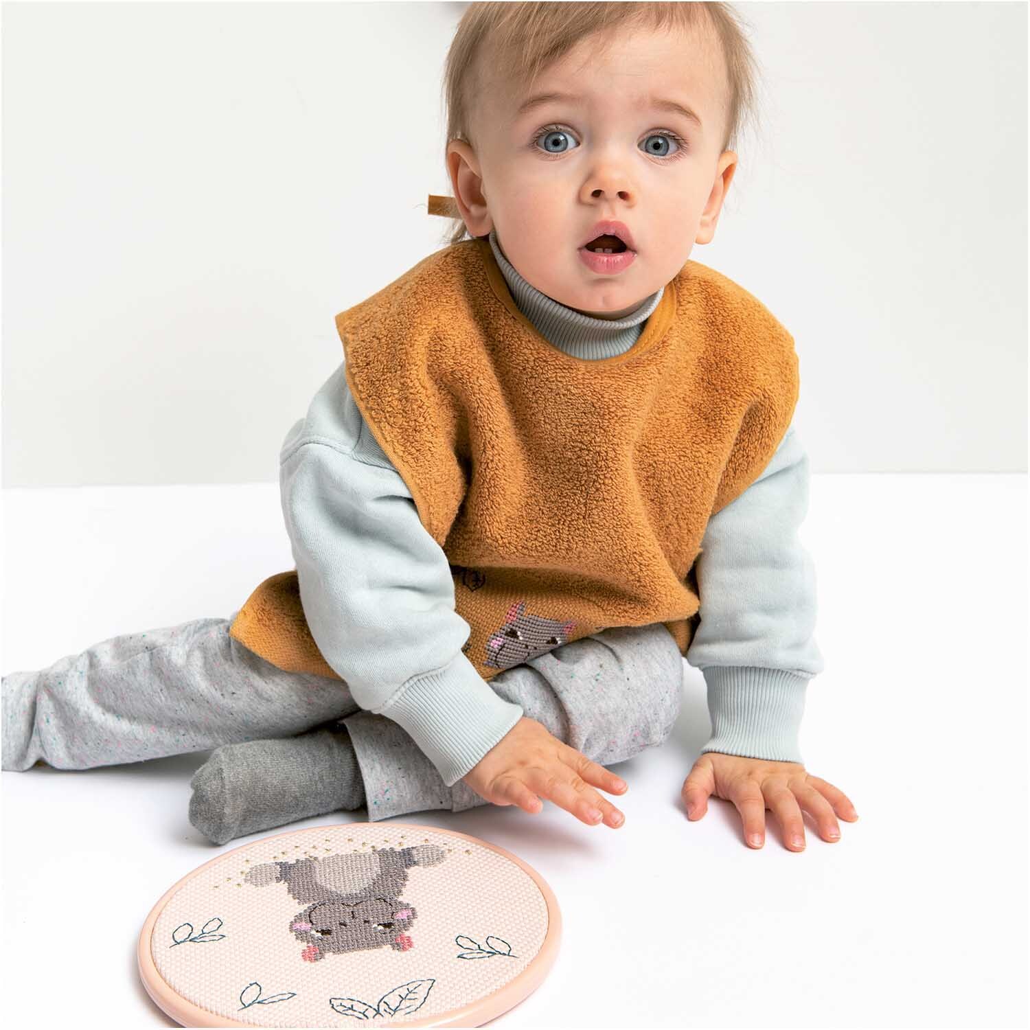 Stickring aus Kunststoff mauve 20,3cm von Rico Design neben einem sitzenden Baby in grauer Kleidung und orangefarbenem Lätzchen.