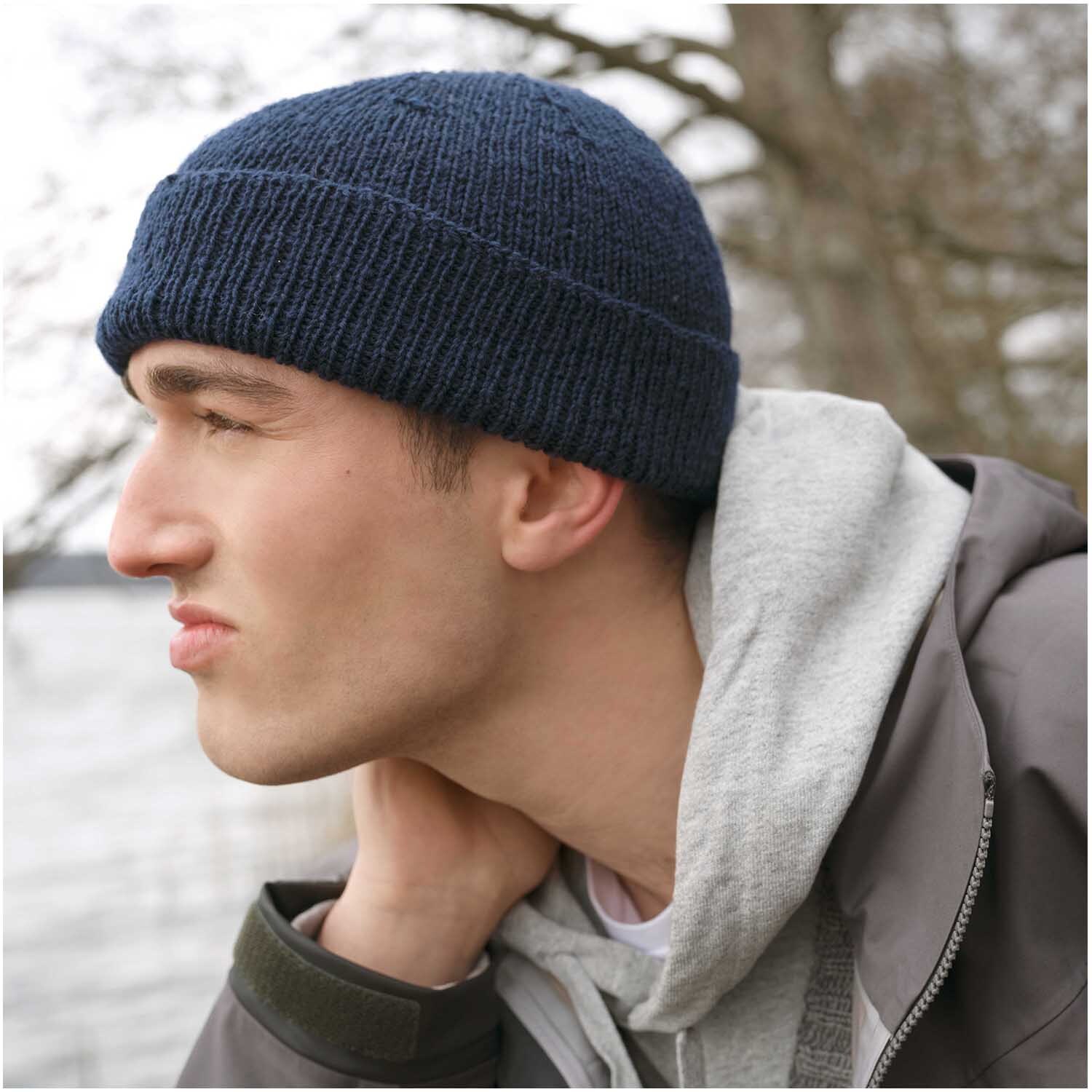Strickset Mütze Modell 09 aus Luxury Knits Winter Special Marine: Mann trägt dunkelblaue Strickmütze im Freien, gekleidet in grauem Hoodie und Jacke.
