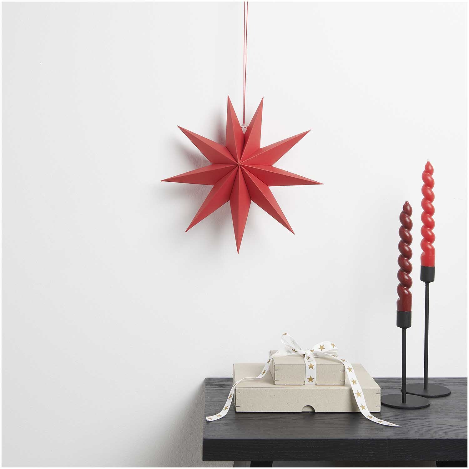 Papierstern auffaltbar 28cm von Rico Design in Rot, dekorativ an der Wand hängend, neben Geschenkbox und gedrehten Kerzen.