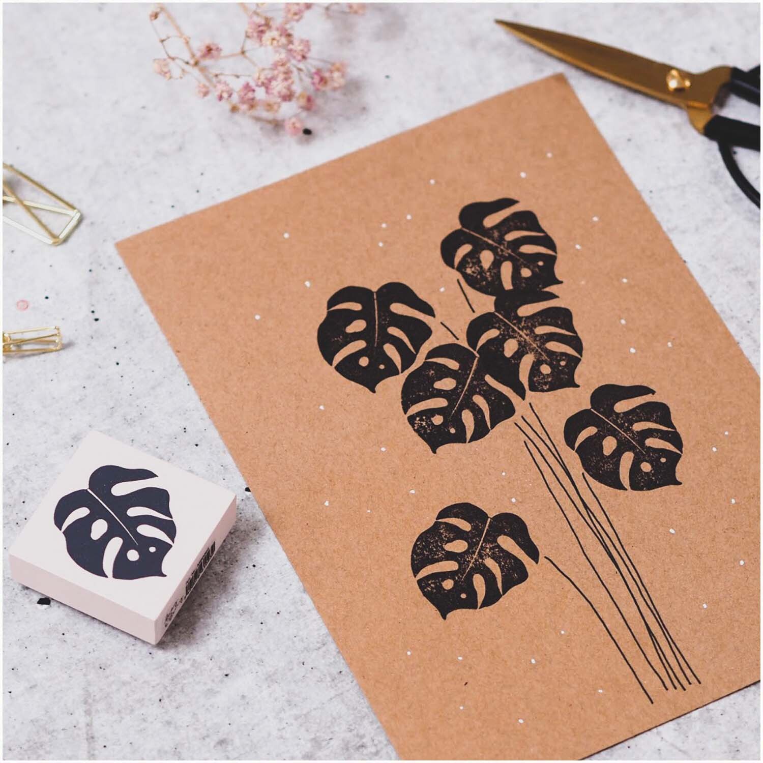 Stempel Palmblatt nude 45x45mm zeigt ein Monstera-Motiv auf braunem Papier, daneben Schere und Büroklammern.
