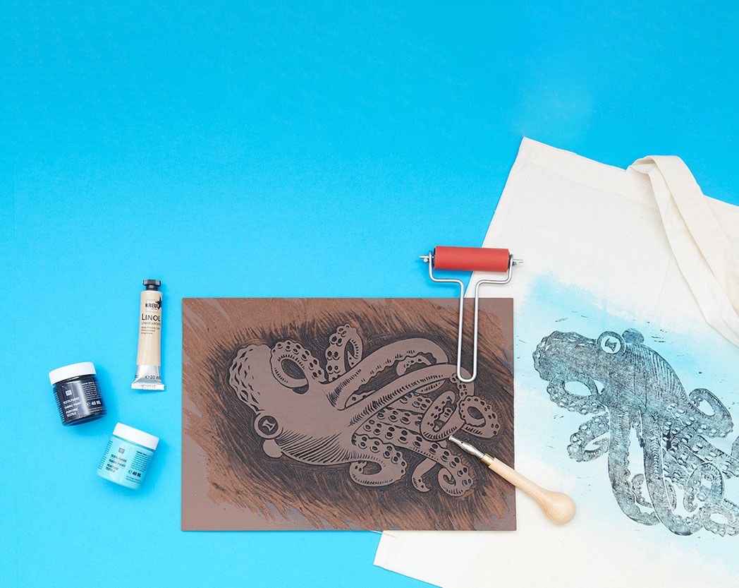 Eine Linolplatte mit einem Oktopus-Motiv hereingeschnitzt, sowie Farben, eine Linolwalze, ein Schneide-Werkzeug und eine bedruckte Tasche. Flatlay von Produkten.