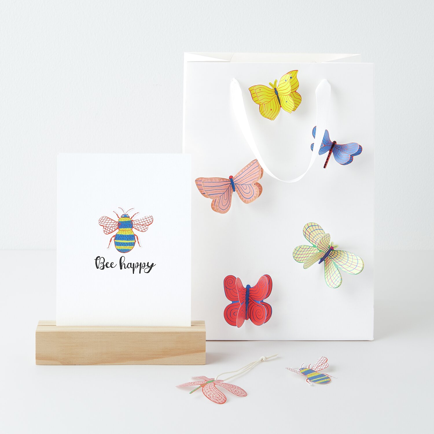 Sticker Midsommar Magic Insekten: Bunte Schmetterlingssticker auf weißer Tasche, daneben Karte mit Biene und "Bee happy"-Text.